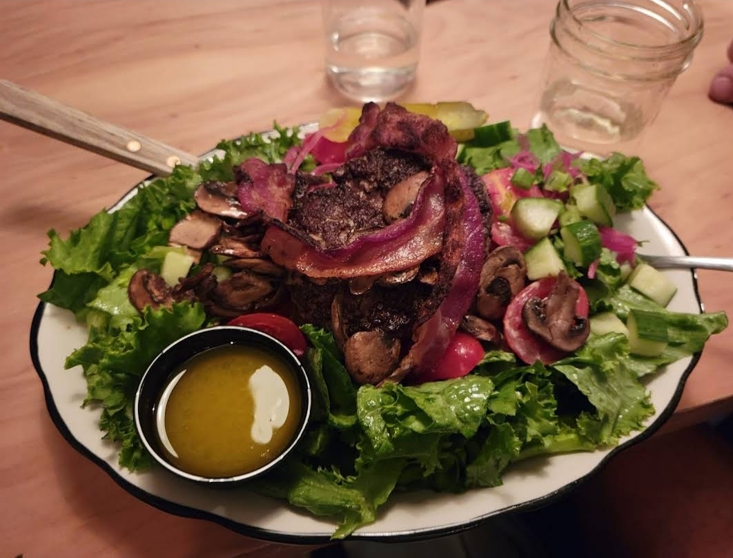 A close-up view of a vibrant salad from Black Tap Craft Burgers & Beer in SoHo, featuring a juicy burger patty topped with crispy bacon, sautéed mushrooms, and red onions, served on a bed of fresh greens with a side of house dressing