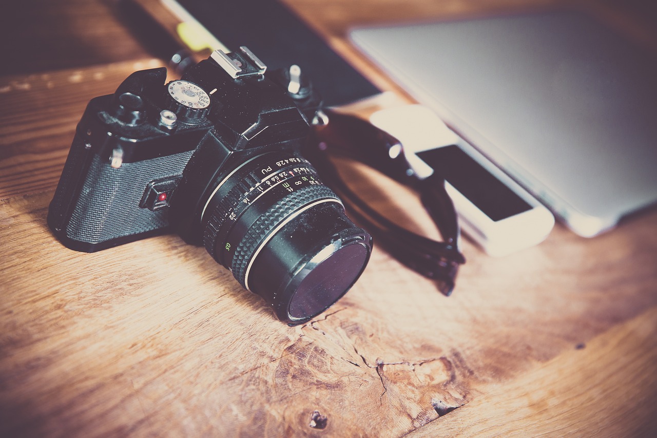 A vintage camera placed on a wooden surface next to a closed laptop and a smartphone.